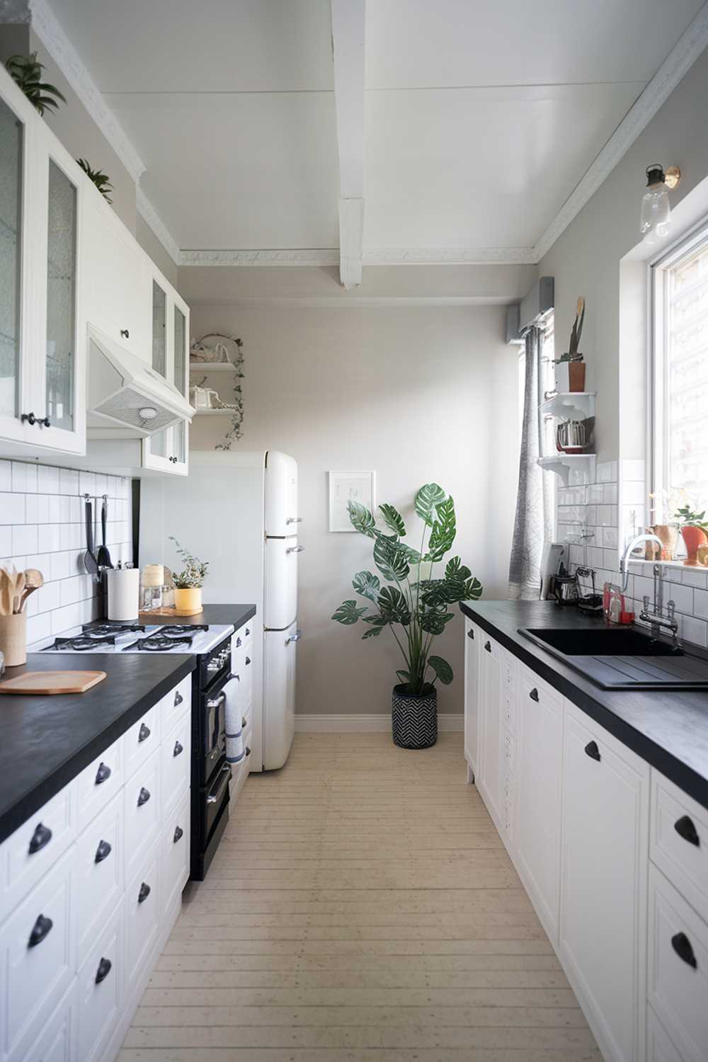 A modern Scandinavian kitchen design. The kitchen features white cabinets, a black countertop, a black stove, a white refrigerator, and a black sink. There is a potted plant near the refrigerator. The floor is made of light wood. The walls are painted in a light gray color. There are a few kitchen tools and utensils on the countertop. The room has a window with a curtain.