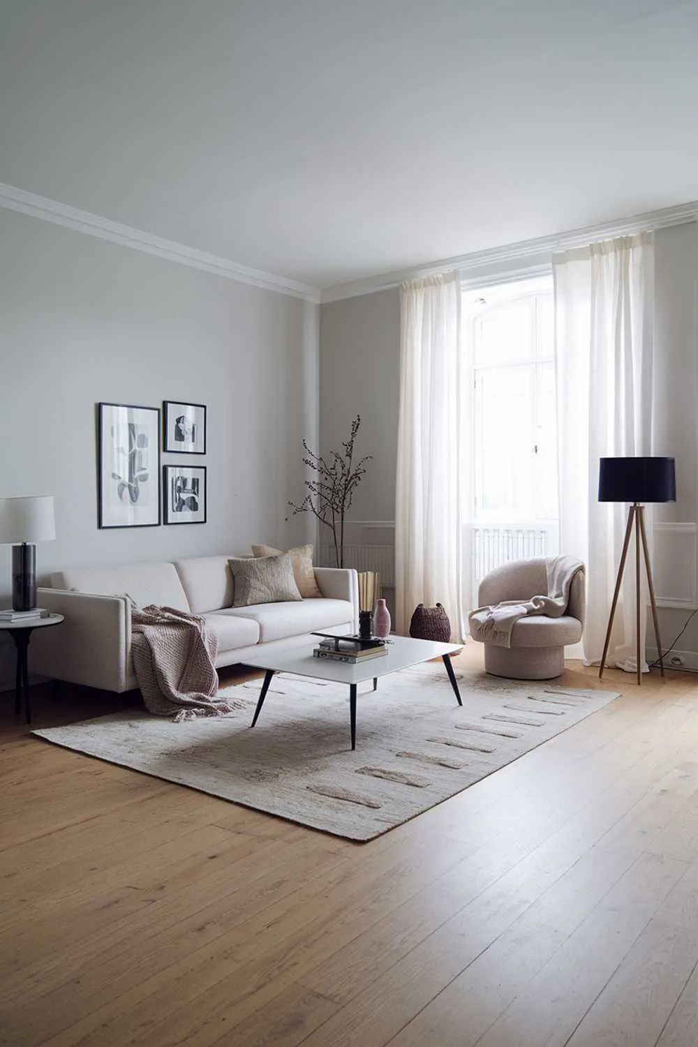 47 Modern Scandinavian Living Room Ideas to Try 1 A modern Scandinavian living room with a light wood floor and a beige sofa. The room has a white coffee table, a black lamp, and a few decorative items. There is a rug in the middle of the room. The walls are painted in light gray. There is a large window with a white curtain.