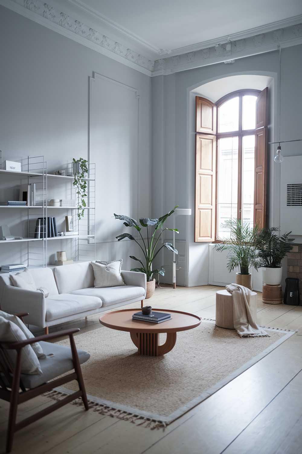 A modern Scandinavian living room with a sleek, minimalist design. The room features a white sofa with a few cushions, a beige rug, a wooden coffee table with a circular design, and a few potted plants. There's a white bookshelf with a few books and decorative items. The walls are painted gray, and there's a large window with wooden shutters. The flooring is light wood.