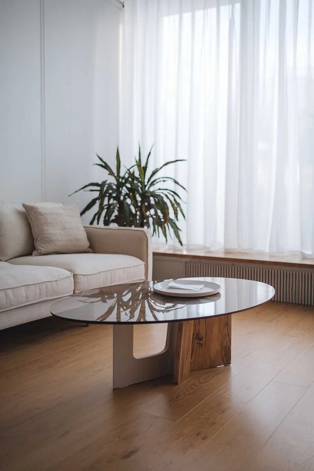 47 Modern Scandinavian Living Room Ideas to Try 6 A close-up shot of a modern Scandinavian living room decor. There's a beige sofa with a few cushions and a coffee table with a wooden base and a glass top. There's a potted plant in the corner. The room has a wooden floor and white walls. There's a large window with white curtains. The light fills the room.