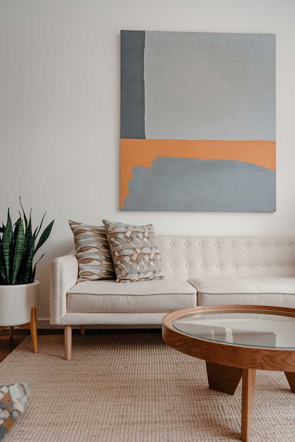 A modern Scandinavian living room decor. There's a beige sofa with a few cushions in a patterned fabric. Next to the sofa is a wooden coffee table with a round, glass top. On the wall behind the sofa is a large, abstract painting with a gray, blue, and orange color palette. The floor is covered with a beige rug. There's a green plant in a white pot on the left side. The lighting is soft.