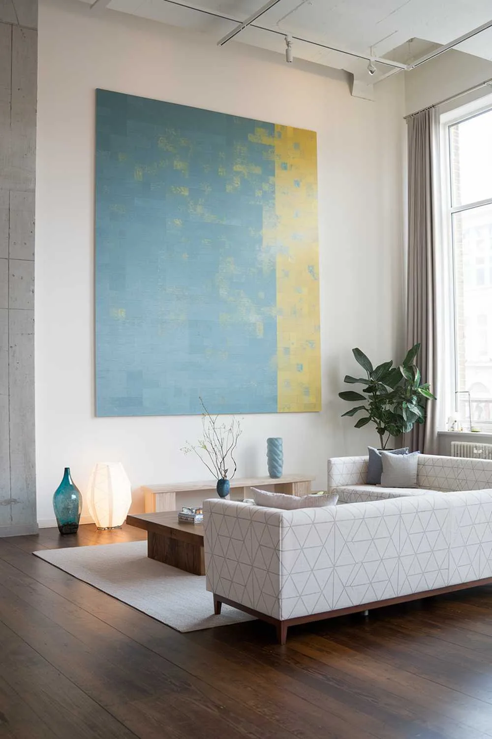 47 Modern Scandinavian Living Room Ideas to Try 19 A large modern Scandinavian living room. The room has a white sofa with a geometric pattern and a few pillows. There's a wooden coffee table in the center. On the wall, there's a large, abstract artwork with shades of blue and yellow. The floor is made of dark wood. There's a potted plant near the window. The room has a few decorative items, such as a small, blue vase and a white, geometric lamp.
