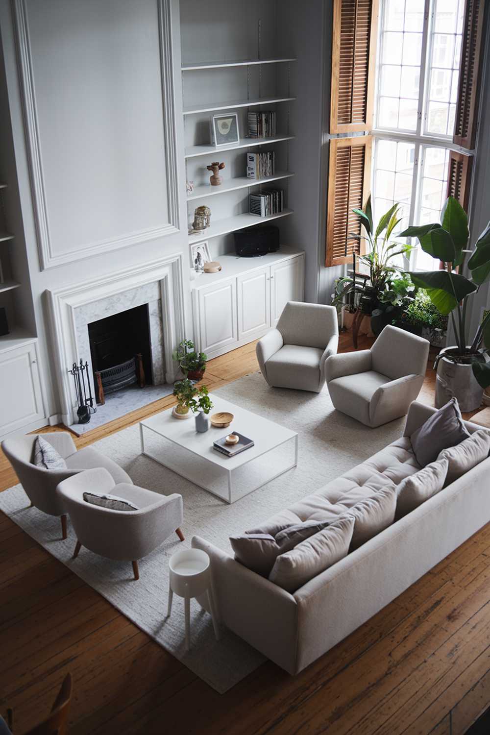 A gray modern Scandinavian living room with a white fireplace. The room has a wooden floor. There's a beige sofa with gray cushions, a few gray chairs, and a white coffee table in the middle of the room. A white bookshelf is placed on the wall behind the sofa. The room has a few plants and a large potted plant near the window. The window has wooden shutters.