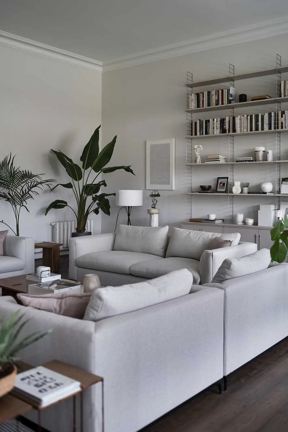 47 Modern Scandinavian Living Room Ideas to Try 25 A large gray modern Scandinavian living room design. The shot is focused on the living room, which has a simple and minimalist design. There are gray sofas with clean lines, a few potted plants, a wooden coffee table, and a white lamp. The walls are painted white, and the floor is made of dark wood. In the background, there's a bookshelf filled with books and decorative items.