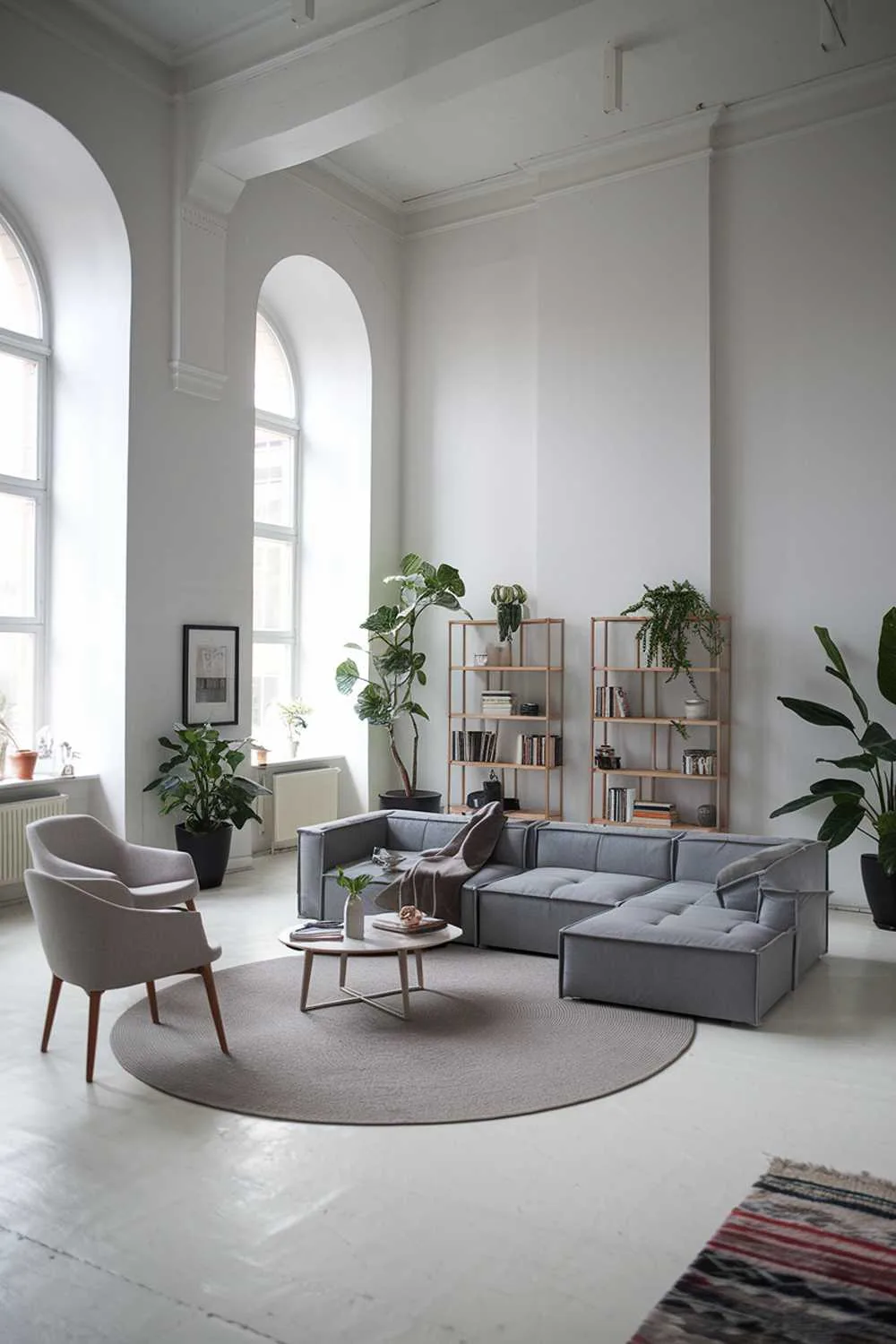 47 Modern Scandinavian Living Room Ideas to Try 26 A large gray modern Scandinavian living room design. The room has a high ceiling and is furnished with a large gray sofa, a few chairs, a coffee table, and a bookshelf. There is a round rug in the center of the room. The walls are white, and there are a few potted plants.