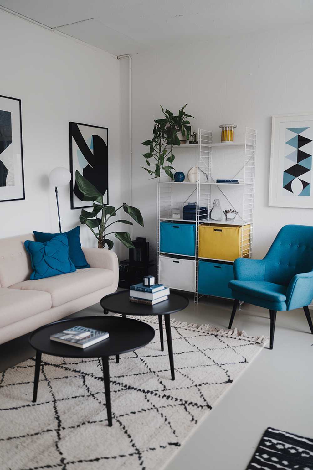 A black and white modern Scandinavian living room with a few pops of color. There's a beige sofa with blue cushions, a black coffee table, a white bookshelf with blue and yellow items, and a blue chair. The room has a rug, a potted plant, and a few decorative items. The walls have a few pieces of art.