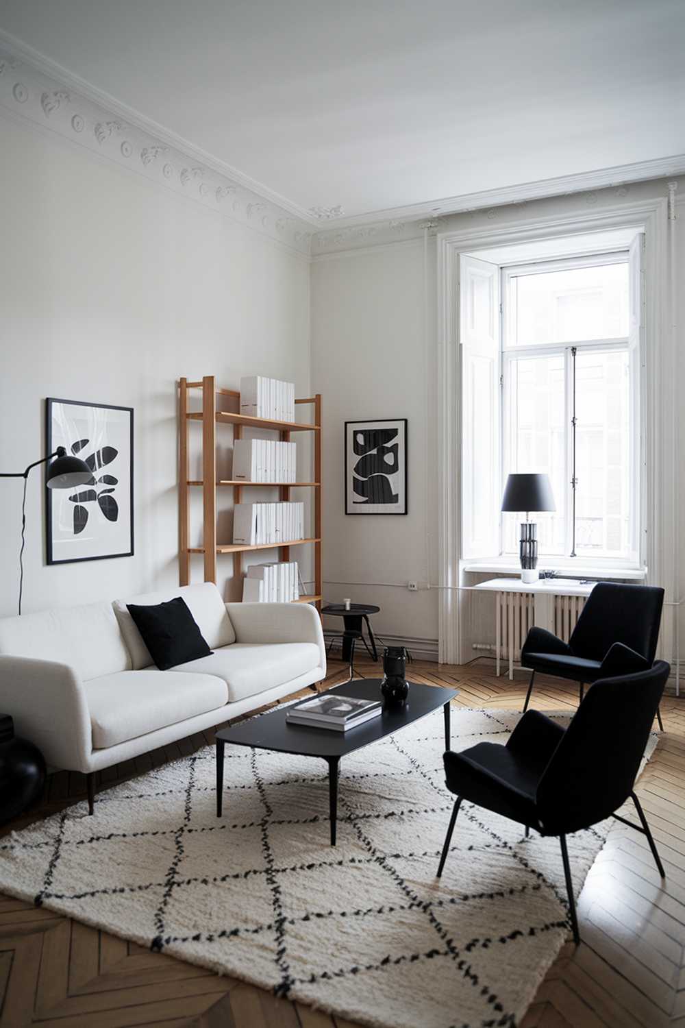 A black and white modern Scandinavian living room design and decor. The room features a white sofa with black cushions, a black coffee table, and a white rug. There are two black chairs near the window. A wooden bookshelf with white books is placed near the sofa. The walls are painted white, and there is a black lampshade on a white table near the window. The room has a wooden floor.