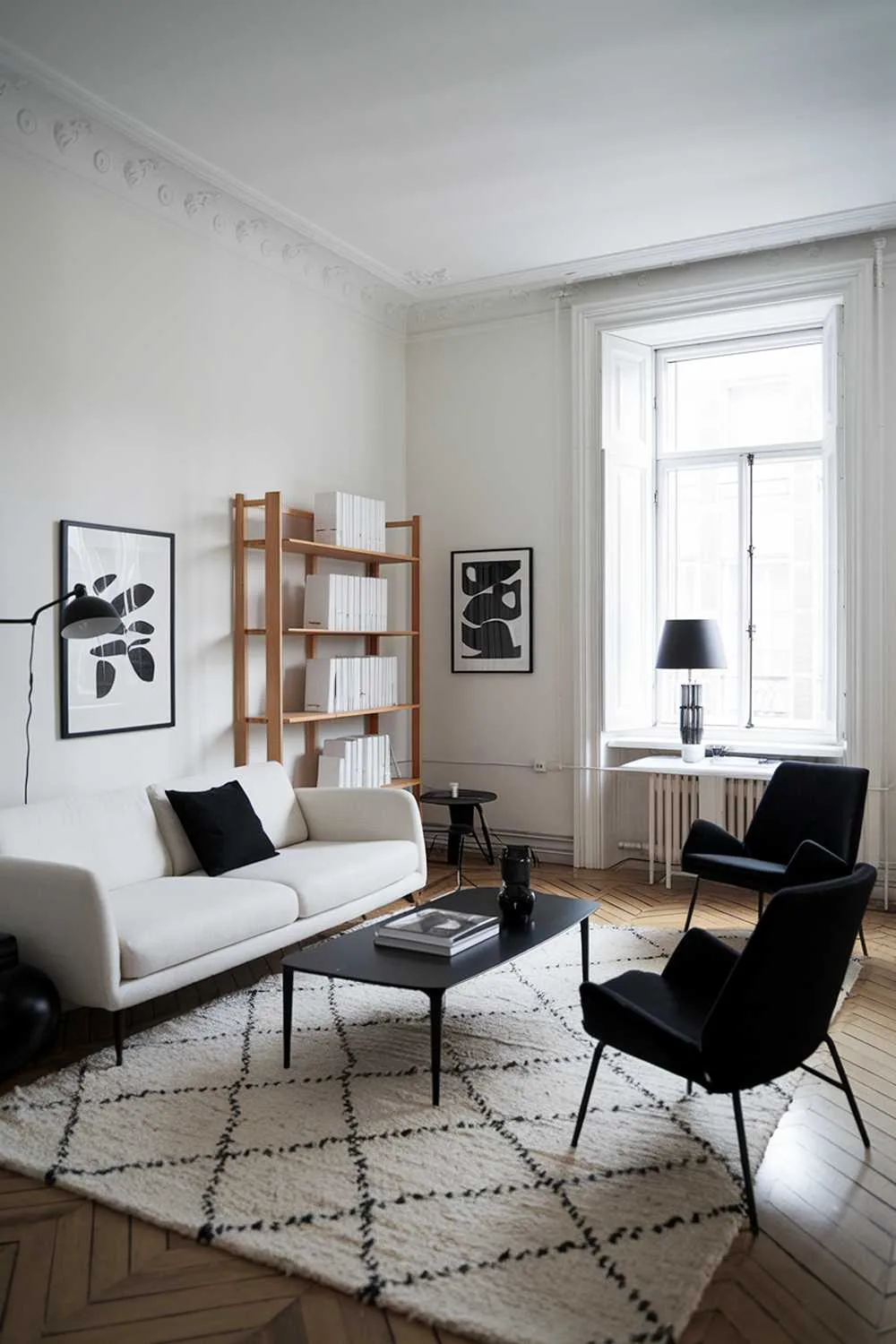 47 Modern Scandinavian Living Room Ideas to Try 29 A black and white modern Scandinavian living room design and decor. The room features a white sofa with black cushions, a black coffee table, and a white rug. There are two black chairs near the window. A wooden bookshelf with white books is placed near the sofa. The walls are painted white, and there is a black lampshade on a white table near the window. The room has a wooden floor.