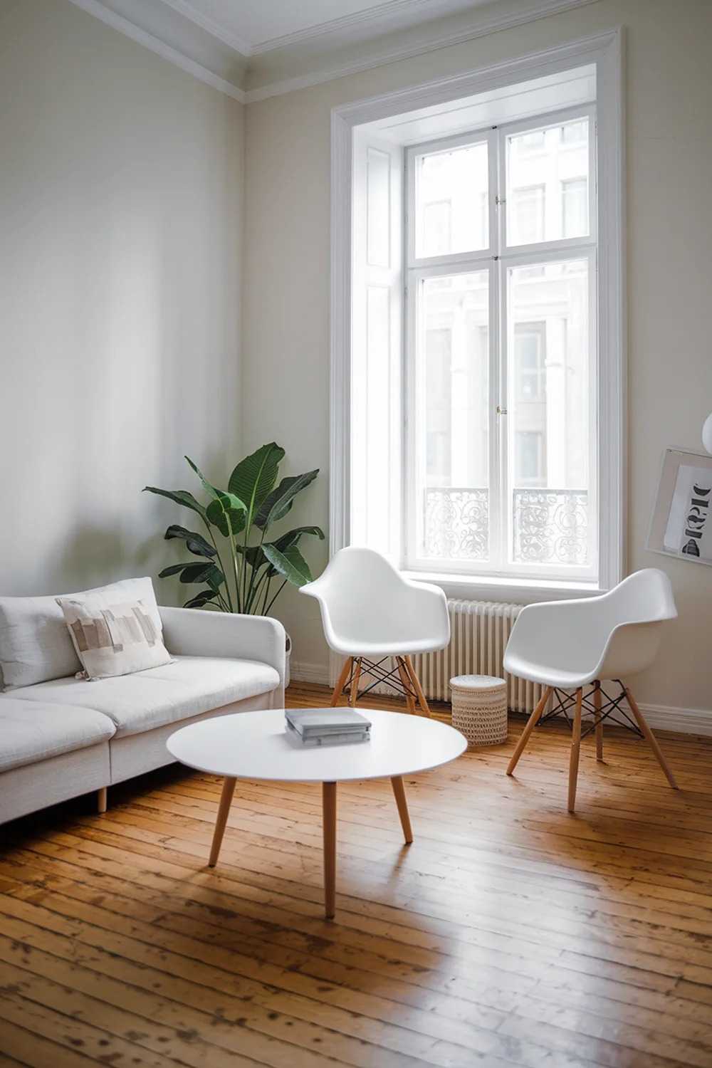 A modern Scandinavian living room with a large window. The room has a white sofa with a few pillows and a round white coffee table. There are two white chairs with black legs near the window. The floor is made of wooden planks. The walls are painted white. There is a green plant near the window.