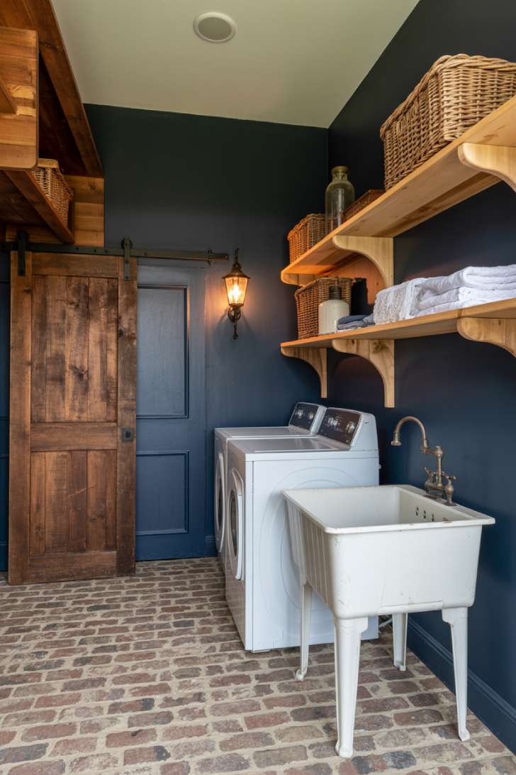 Moody farmhouse laundry room with navy walls, brick floor, reclaimed wood shelves, and cast iron sink
