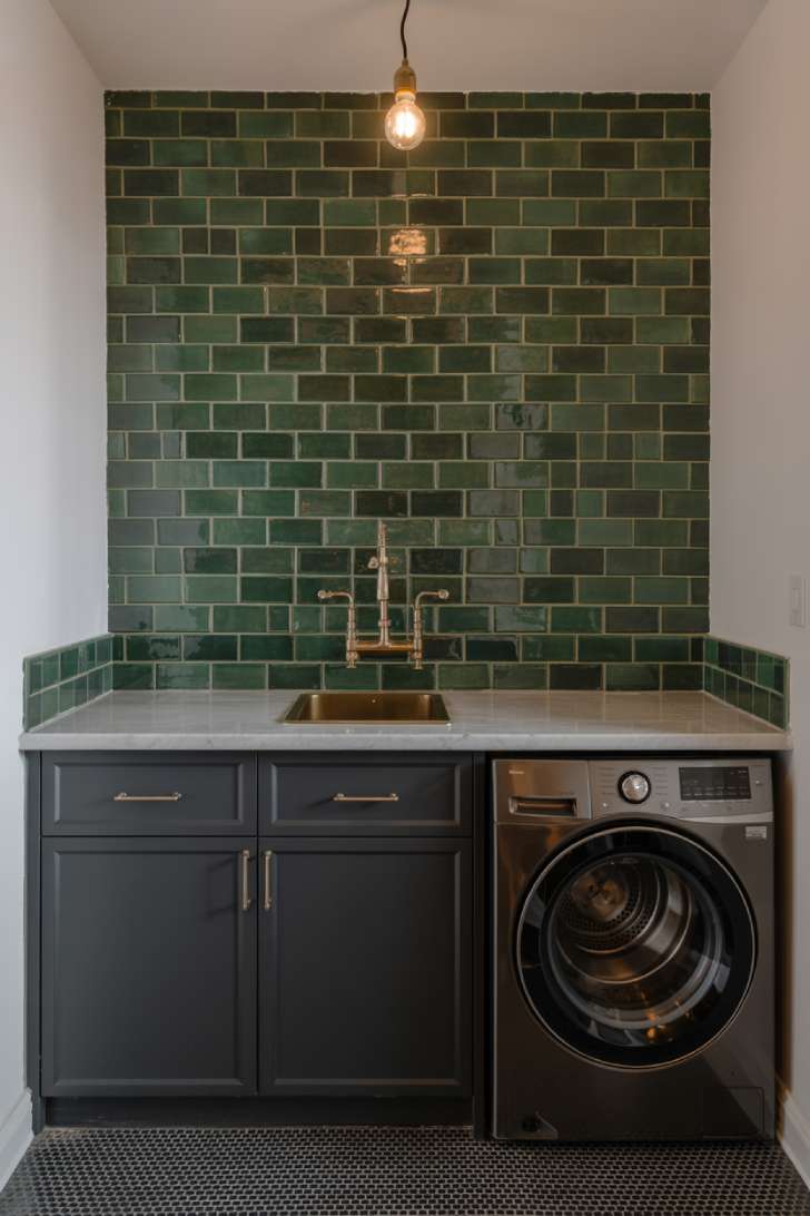 Forest green subway tile laundry room wall with black cabinets, marble countertop, and brass fixtures