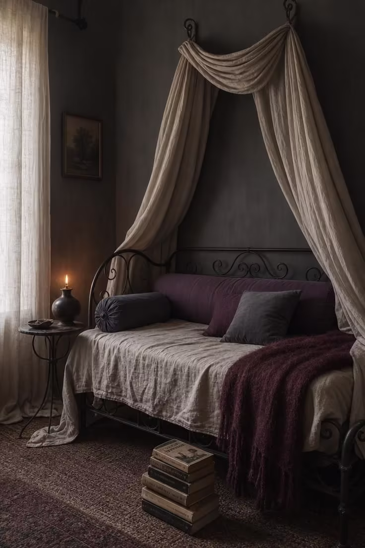 14 Moody Reading Nook Ideas That Feel Like a Secret Escape 2 A wrought iron daybed with an unbleached linen canopy, plum and slate bolster pillows, and a ceramic oil lamp on a low iron side table in soft diffused light.
