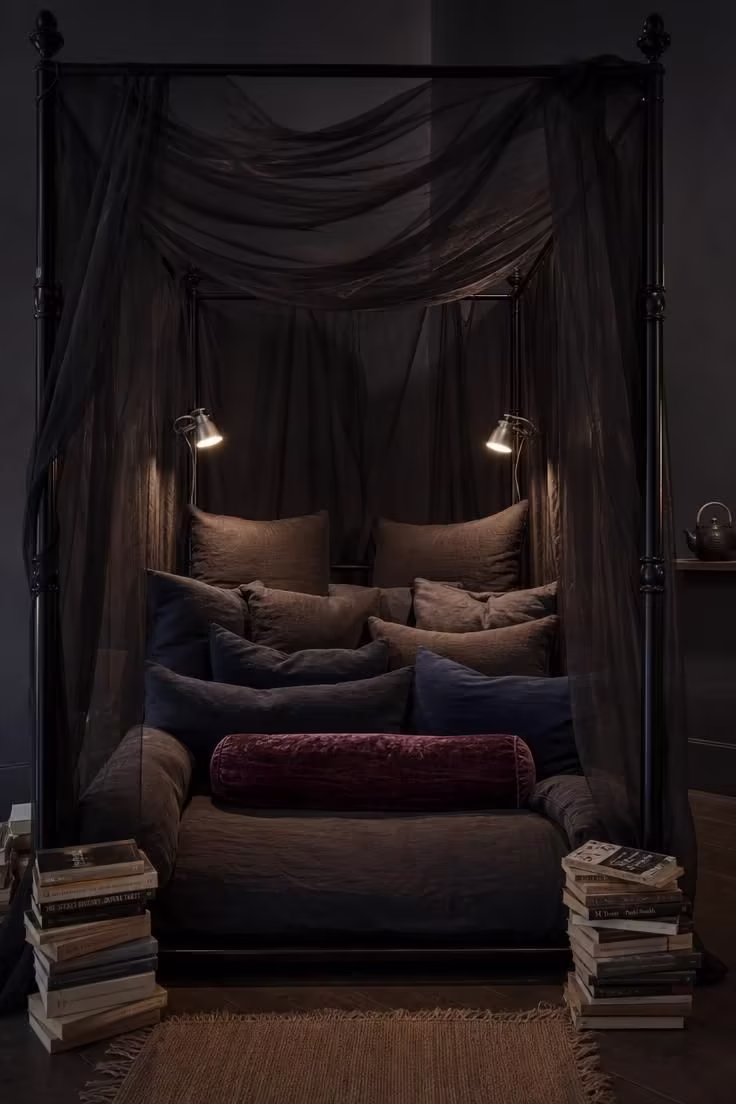 14 Moody Reading Nook Ideas That Feel Like a Secret Escape 11 A four-poster iron canopy bed frame repurposed as a reading lounge with layered charcoal and navy cushions, sheer dark organza canopy panels, clip-on reading lights, and book stacks on the floor.