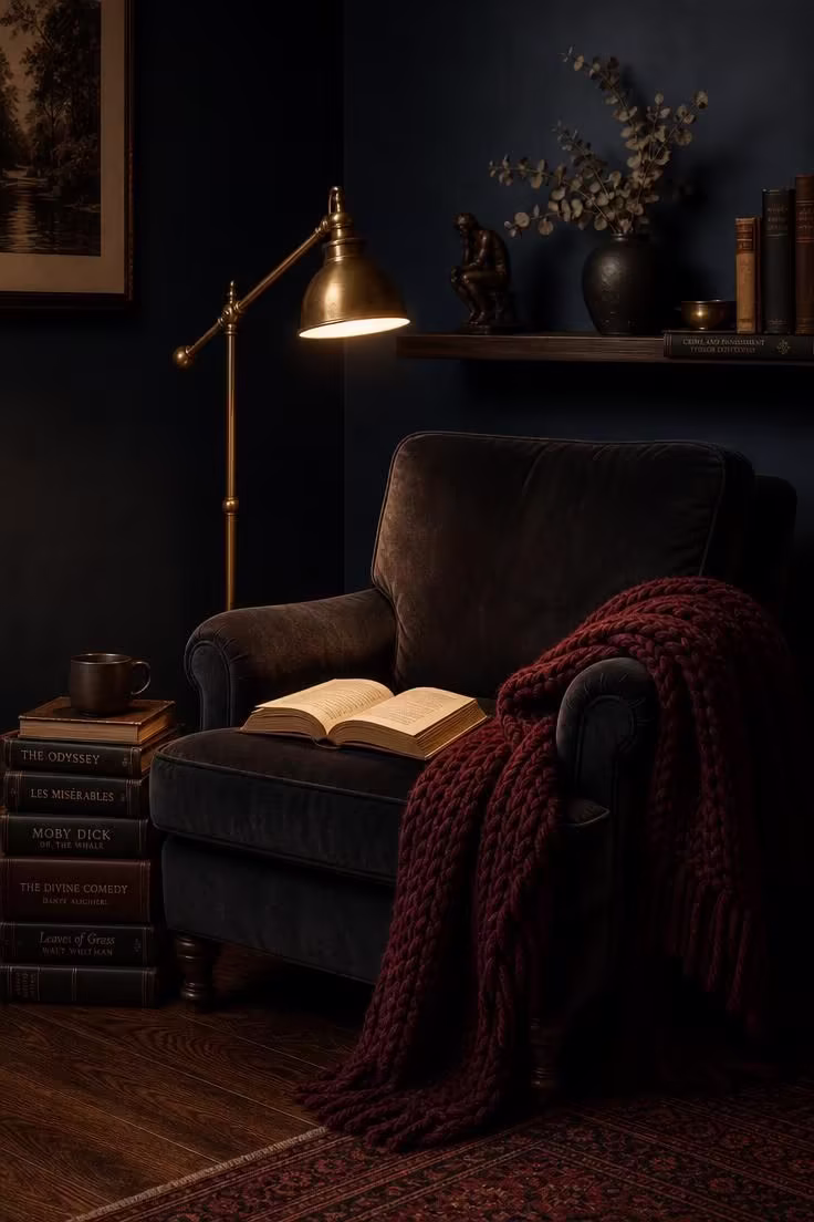 Simple Spaces 14 A moody reading nook with a deep charcoal velvet armchair, brass pharmacy lamp, chunky burgundy throw, hardcover books on a dark walnut floor, and a midnight blue wall with a floating shelf of collected objects.
