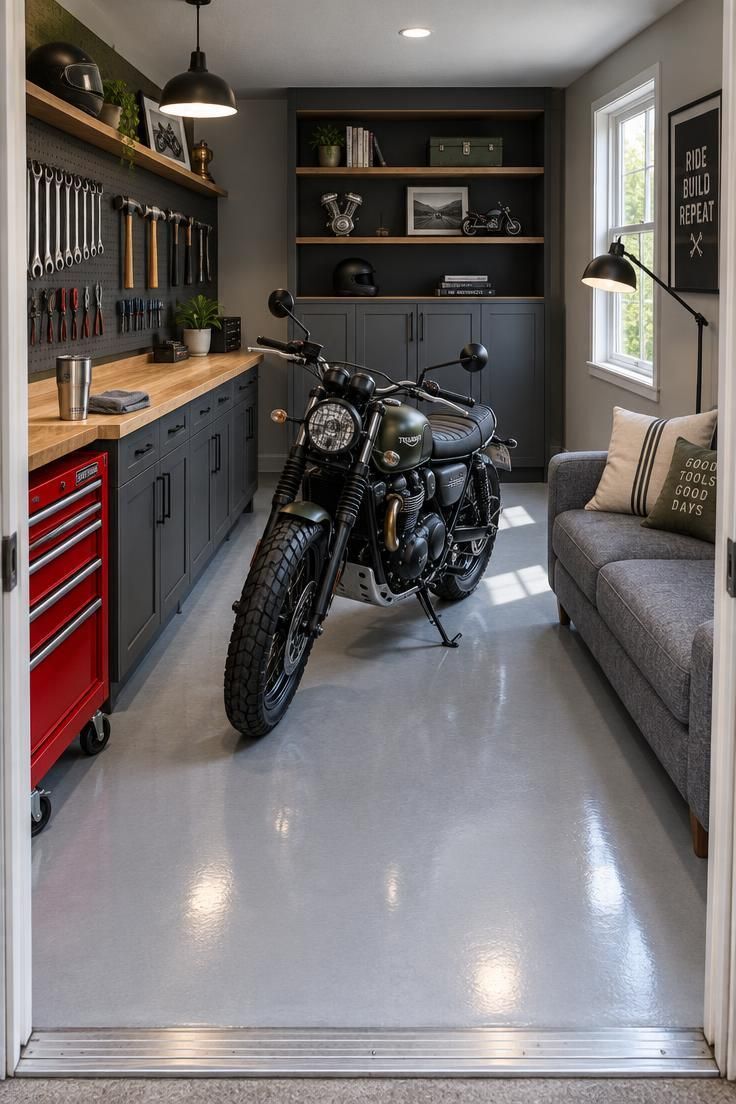 10 Cool Motorcycle Man Cave Ideas That Feel Like a Ride 3 A garage with a wooden workbench and red tool chest on one side, a gray sofa on the other, and a motorcycle angled in the center on a light gray epoxy floor.