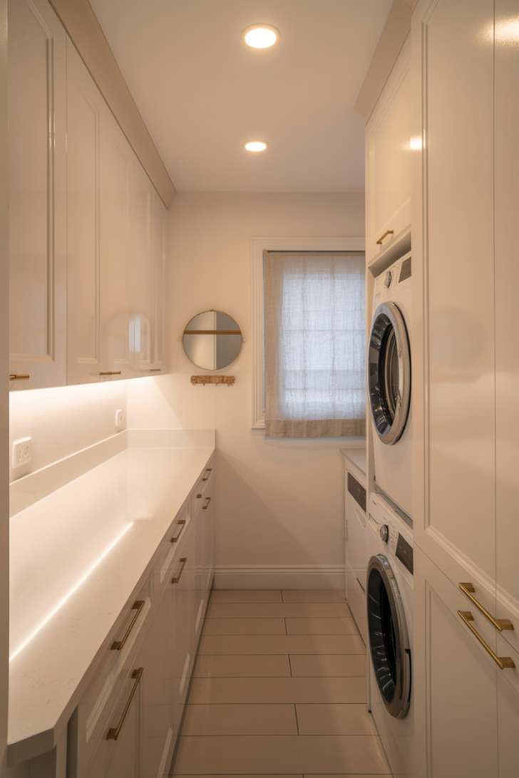 A bright white narrow laundry room with under-cabinet lighting, a brass mirror, and a small sheer-curtained window