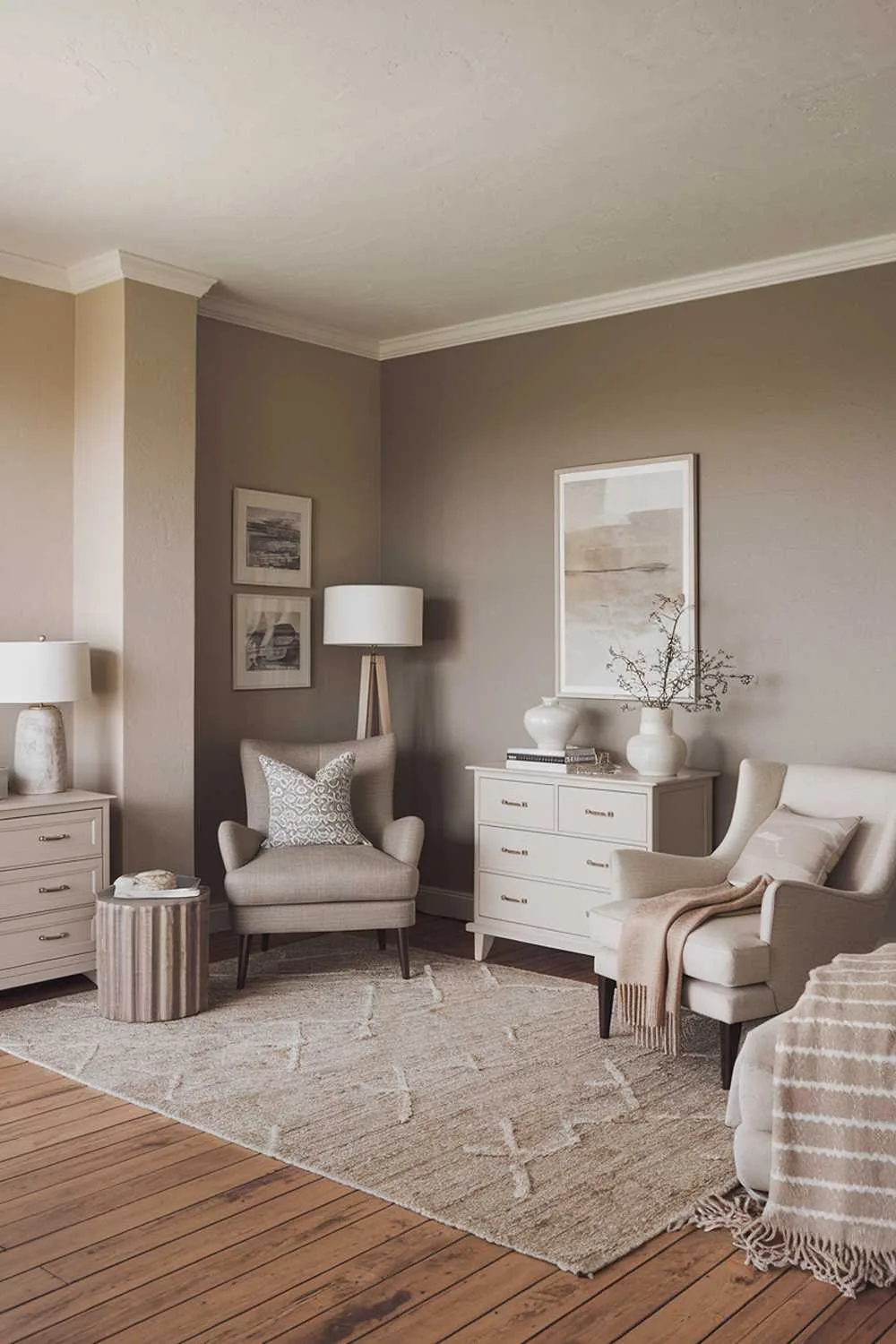 A cozy, neutral bedroom with a high-quality texture. The room features a wooden floor, a beige area rug, a white dresser, a gray chair, a beige armchair, a white lamp, a beige wall, and a gray wall. There is a patterned throw pillow on the gray chair and a beige throw blanket on the beige armchair. The room has soft lighting.