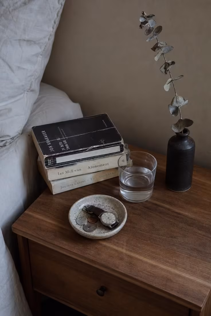 Overhead view of a walnut nightstand with well-read books, a handmade ceramic dish, and a stoneware bottle on a cool overcast morning.
