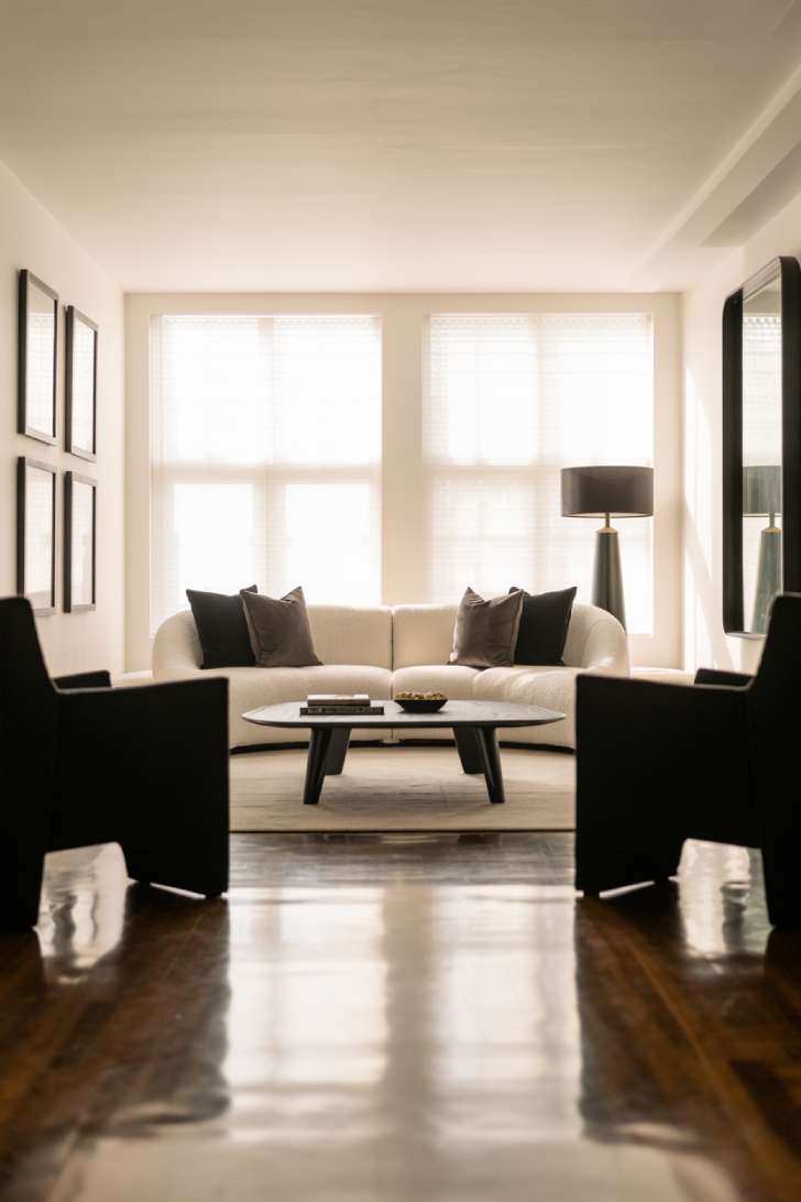 Spacious neutral living room with cream sectional sofa, black geometric coffee table, black picture frames, and a black drum-shade floor lamp on polished hardwood floors