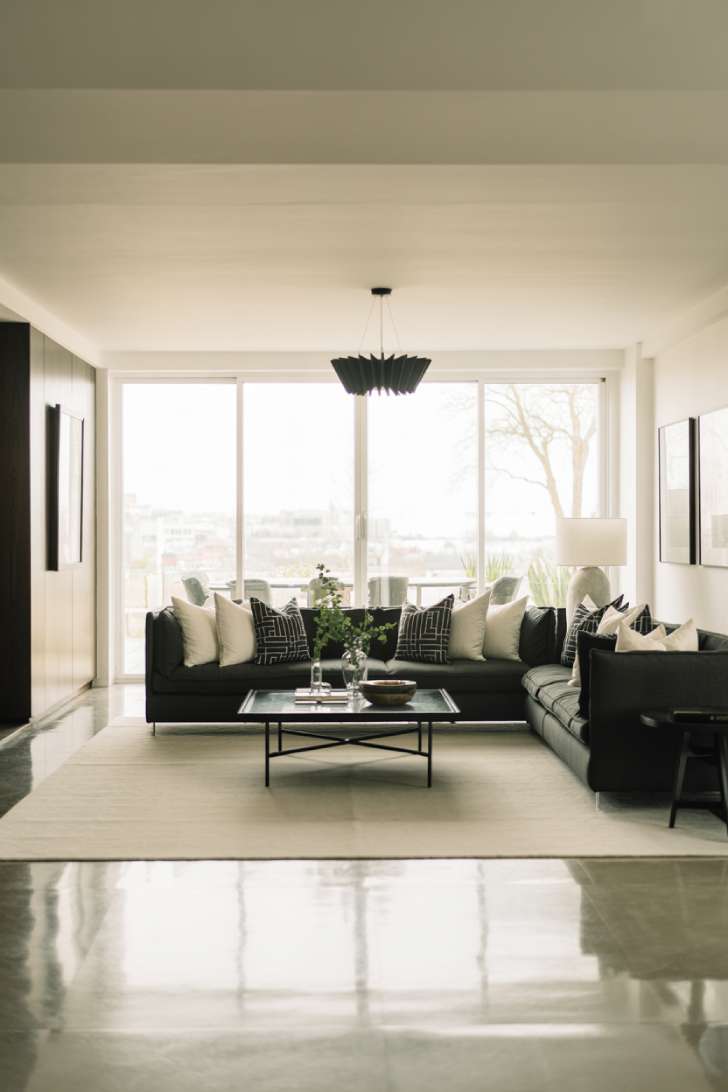 Charcoal gray sectional sofa with white and black geometric pillows, black coffee table with metal legs, floor-to-ceiling windows, black pendant light, black picture frames, cream area rug, and potted greenery