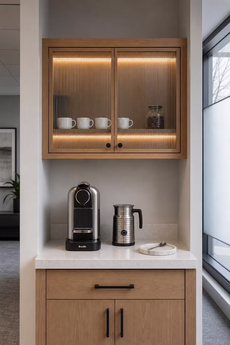10 Cozy Office Coffee Bar Ideas for a Productive Nook 7 Oak cabinet with ribbed glass doors and LED strip lighting above a pod machine on a quartz countertop in a modern office coffee bar