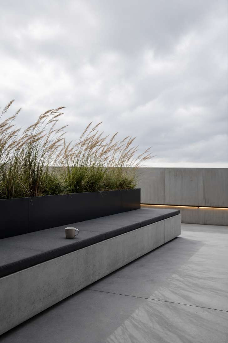 Minimalist open terrace with polished concrete floor, long concrete bench, charcoal cushion, black steel planter with ornamental grasses.