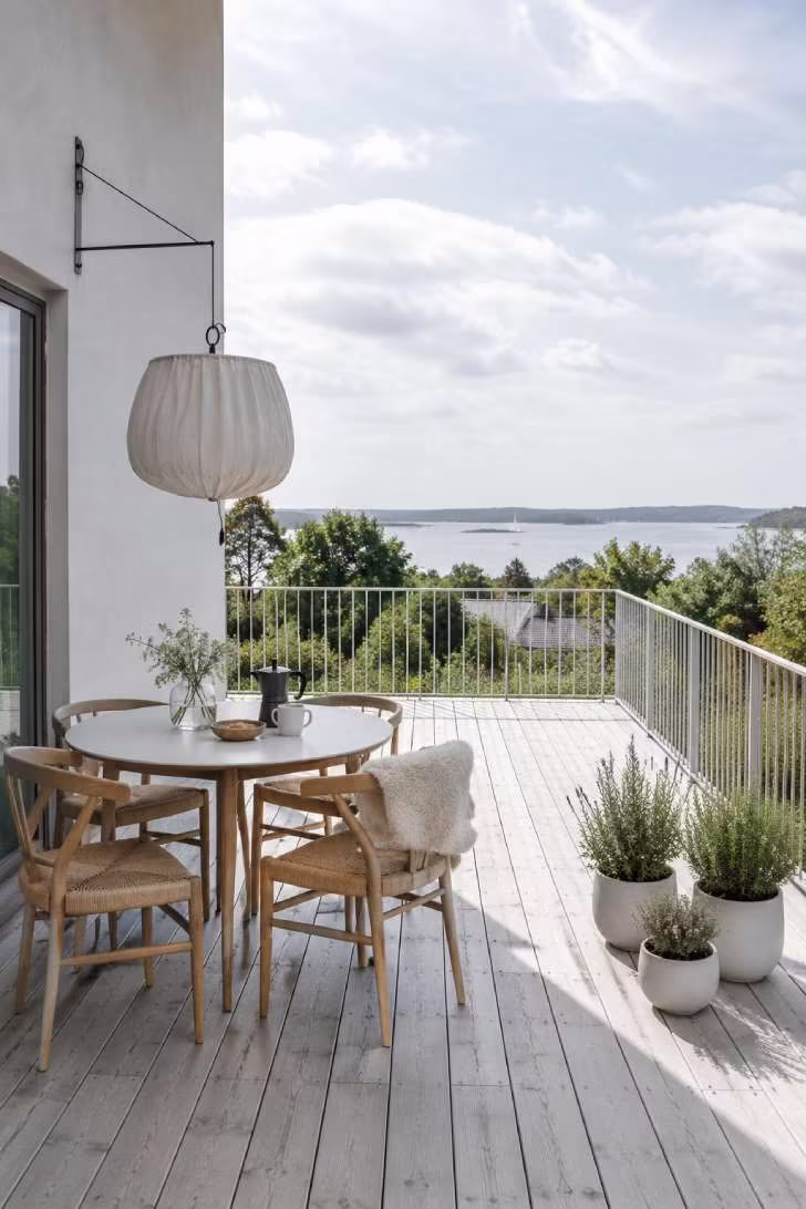 Scandinavian open terrace with pale ash decking, round white table, oak chairs, sheepskin throw, herb pots, and linen pendant shade.