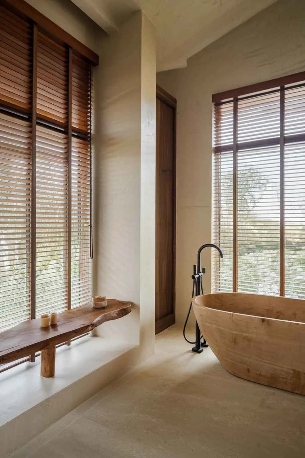26 Stylish Organic Modern Bathroom Design Ideas 3 A minimalist organic modern bathroom with wooden blinds on a large window, freestanding natural material bathtub, simple wooden shelf, beige tiled floor, and visible hardware details.