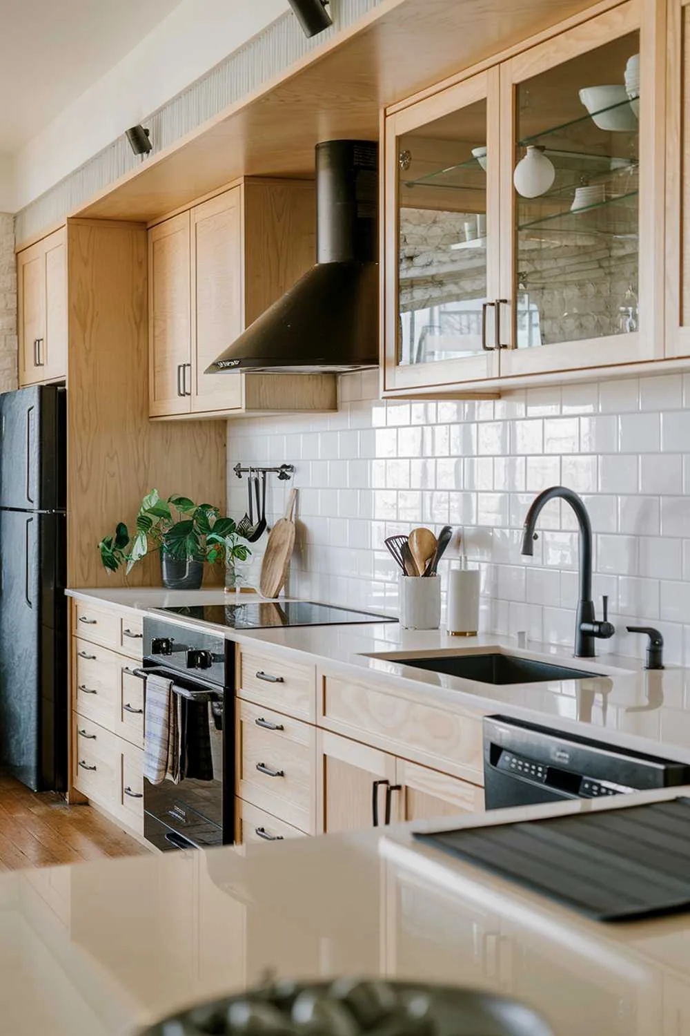 An organic modern kitchen design. The kitchen has a light wood cabinetry, white countertops, and a white backsplash. There is a black sink and a black faucet. There are also a few black appliances, including a refrigerator, a range hood, and a dishwasher. A few utensils are placed on the countertop. There is a green plant near the sink. The floor is made of wood.