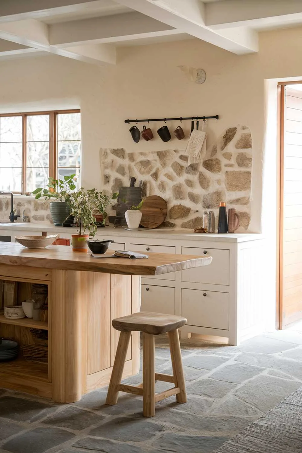 An organic modern kitchen design with a wooden island, white cabinets, and a stone backsplash. The island has a few objects, including a plant and a bowl. There's a wooden stool near the island. The floor is made of gray stone. The wall behind the island has a few hanging pots. The background contains a window with natural light and a door.