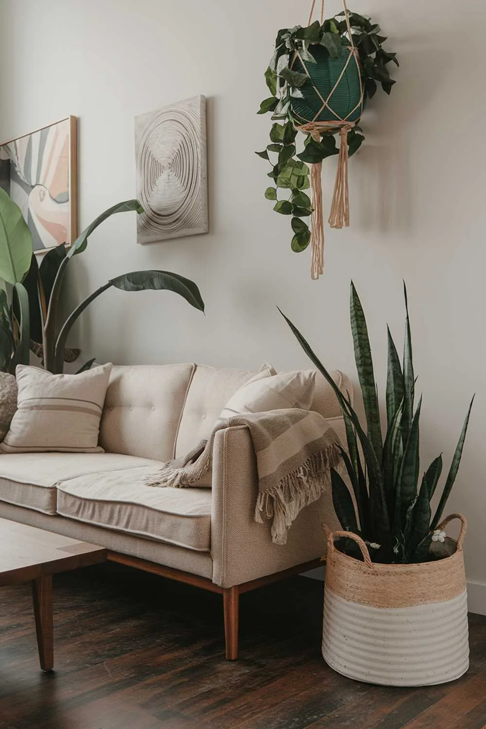 A close up shot of a cozy organic modern living room decor. There is a beige sofa with a few throw pillows, a wooden coffee table, and a large plant. The wall has a few pieces of hanging greenery and a few pieces of wall art. The floor is made of dark wood.