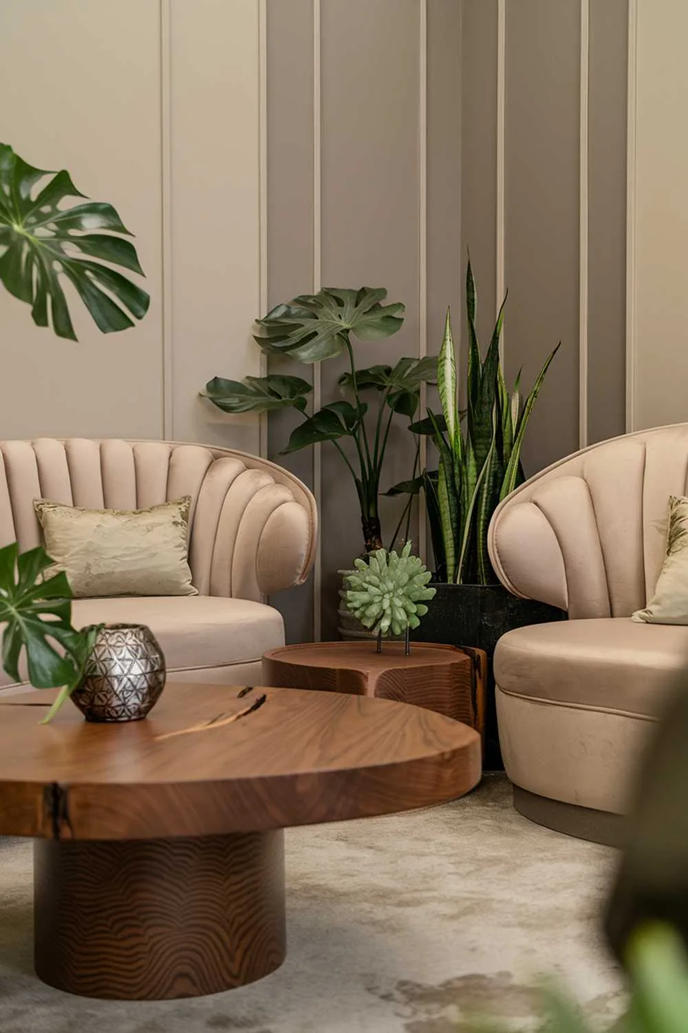 A stylish organic modern living room decor. The room features a beige sofa with a curved backrest and a few cushions. There is a wooden coffee table with a circular base and a unique design. A few potted plants, including a monstera and a snake plant, are placed on the floor and on the table. There is a decorative object with a geometric pattern on the table. The walls have a beige and grey pattern.