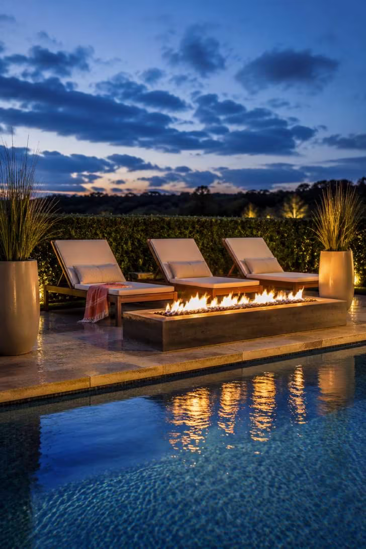 15 Dreamy Outdoor Fire Pit Area Ideas for Every Budget 11 A rectangular dark concrete fire pit between a turquoise pool and teak chaise lounges on travertine coping, reflecting fire and twilight sky in the water.