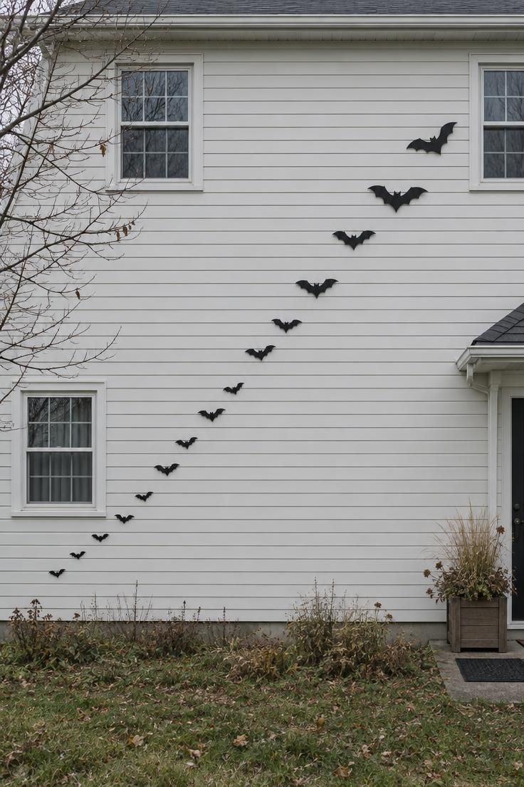 11 Chilling Outdoor Halloween Decor Ideas for a Spooky Night 1 Matte black bat silhouettes rising in a diagonal line across a pale siding wall on an overcast autumn day.