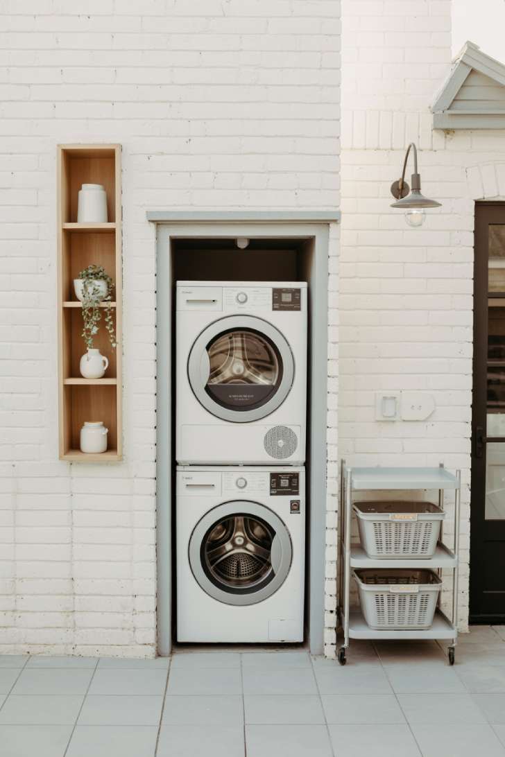 10 Creative Outdoor Laundry Room Ideas for Small Spaces 4 A compact stacked washer and dryer against a white brick wall with oak floating shelves and a pull-out laundry trolley on gray tile