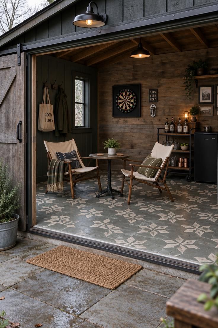 A barn-style outbuilding with a sliding door open wide, a seating group of two chairs and a small table inside, a dartboard and bottle opener on the wall, and a bar cart.