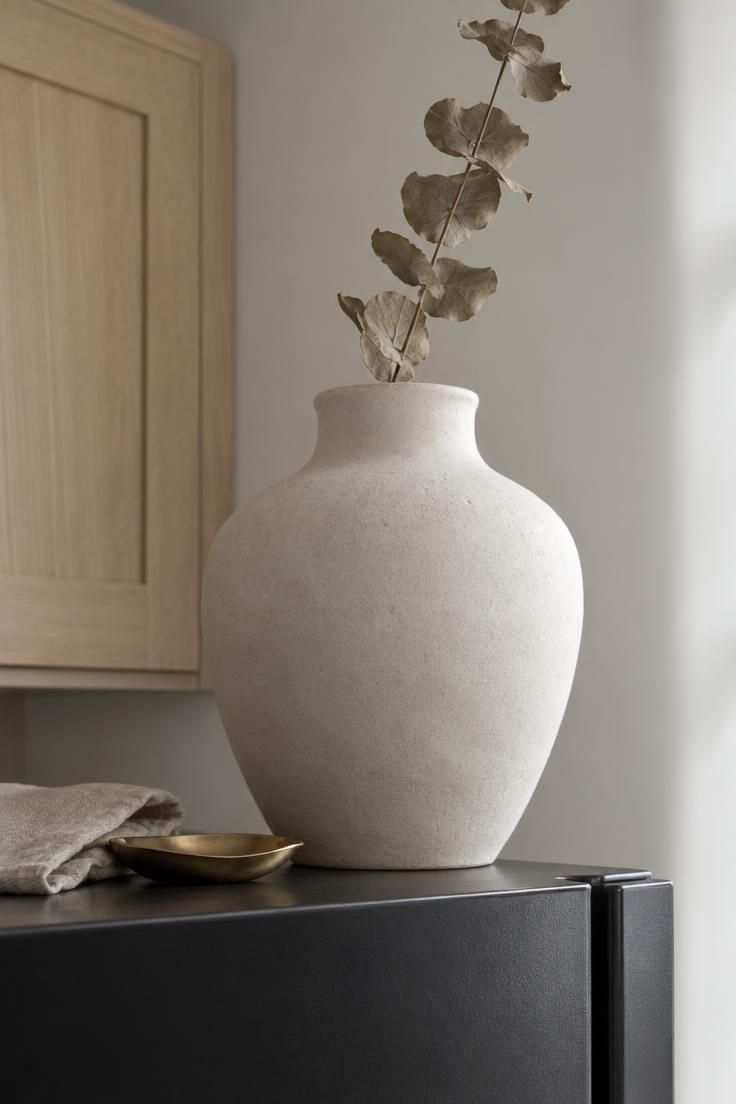 9 Cozy Over the Fridge Decor Ideas for a Warm Kitchen 2 An oversized matte white ceramic urn holding a tall dried branch on a dark refrigerator top with a soft-focus cabinet edge behind it.