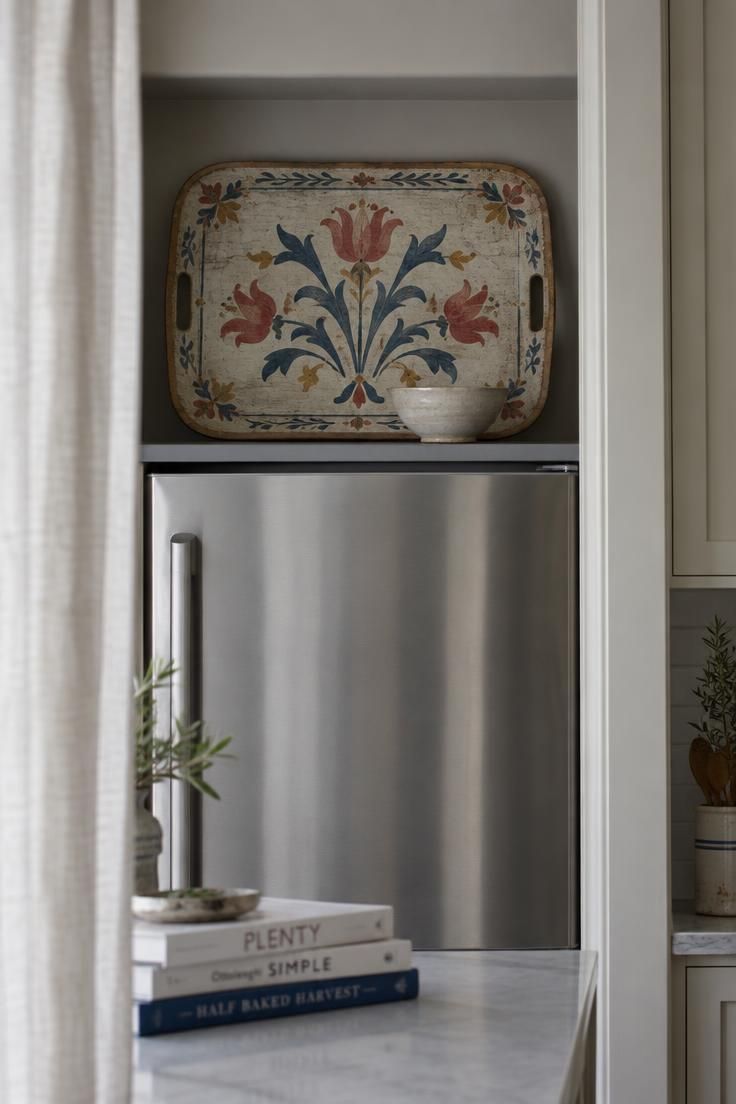 9 Clever Over the Fridge Storage Ideas That End Clutter 5 A large hand-painted folk-motif wooden tray leaning against the back wall above a refrigerator with a small cream bowl in front, cool overcast daylight seen from a kitchen doorway.