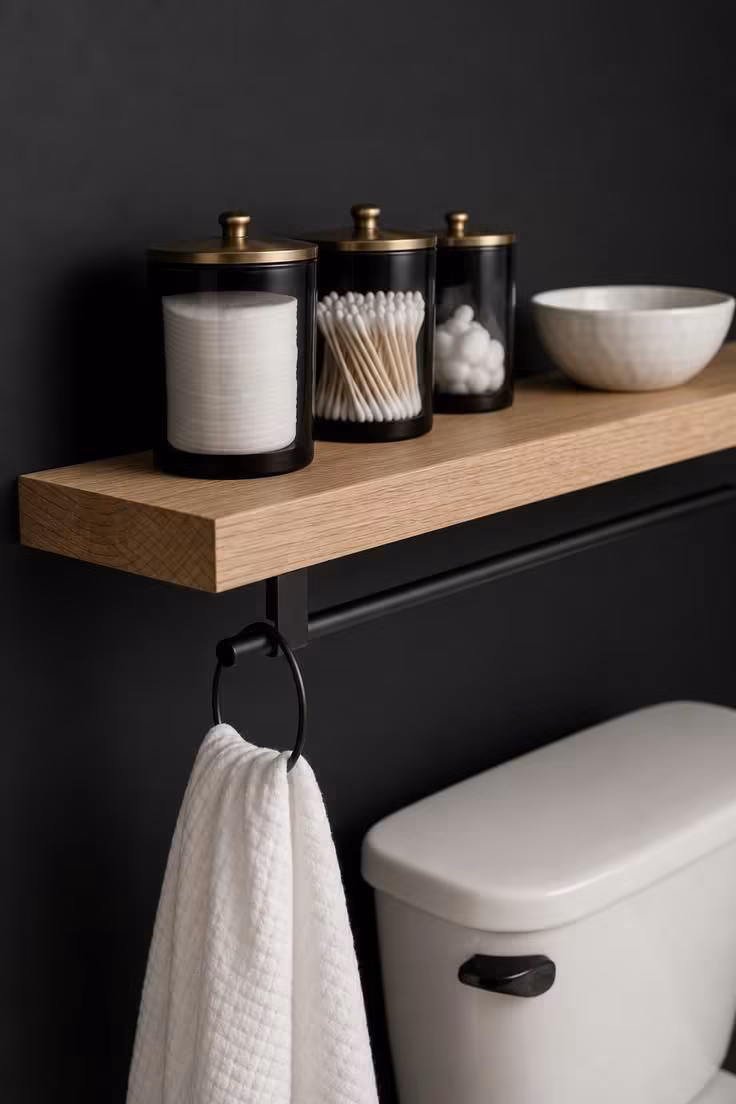 10 Over the Toilet Storage Ideas to End Clutter Now 10 White oak floating shelf over a matte black toilet wall, holding black and brass canisters and a hand towel rail.