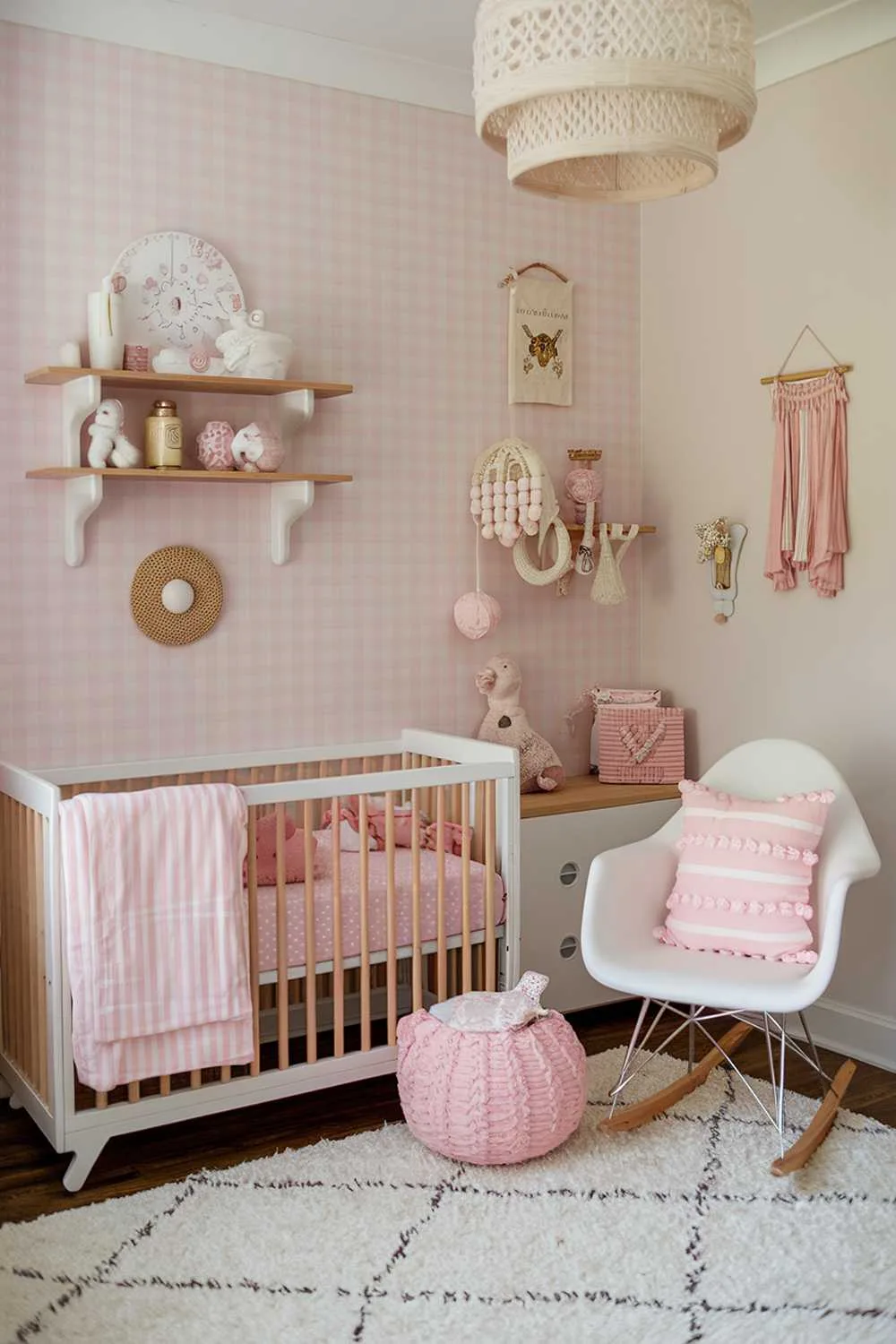 A pink boho nursery room decor. There's a crib with a pink bedding set and a pink and white striped blanket. There's a white rocking chair with a pink cushion. The walls are adorned with a pink and white gingham pattern. There's a wooden shelf with white and pink items. The floor is covered with a white rug.