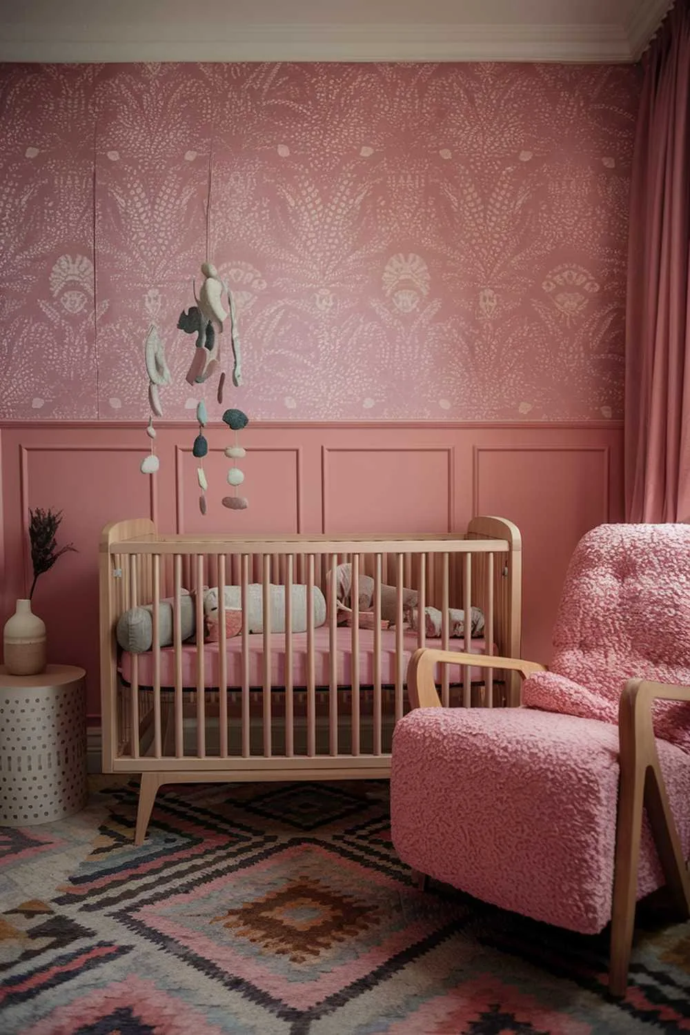 A pink boho nursery room with a cozy and stylish ambiance. The room has a pink wall with a patterned wallpaper. There's a wooden crib with a pink cushion. A plush pink chair with a wooden frame is placed near the crib. A hanging mobile with various shapes is above the crib. The floor is covered with a patterned rug. There's a side table with a decorative vase near the crib. The room has soft lighting.