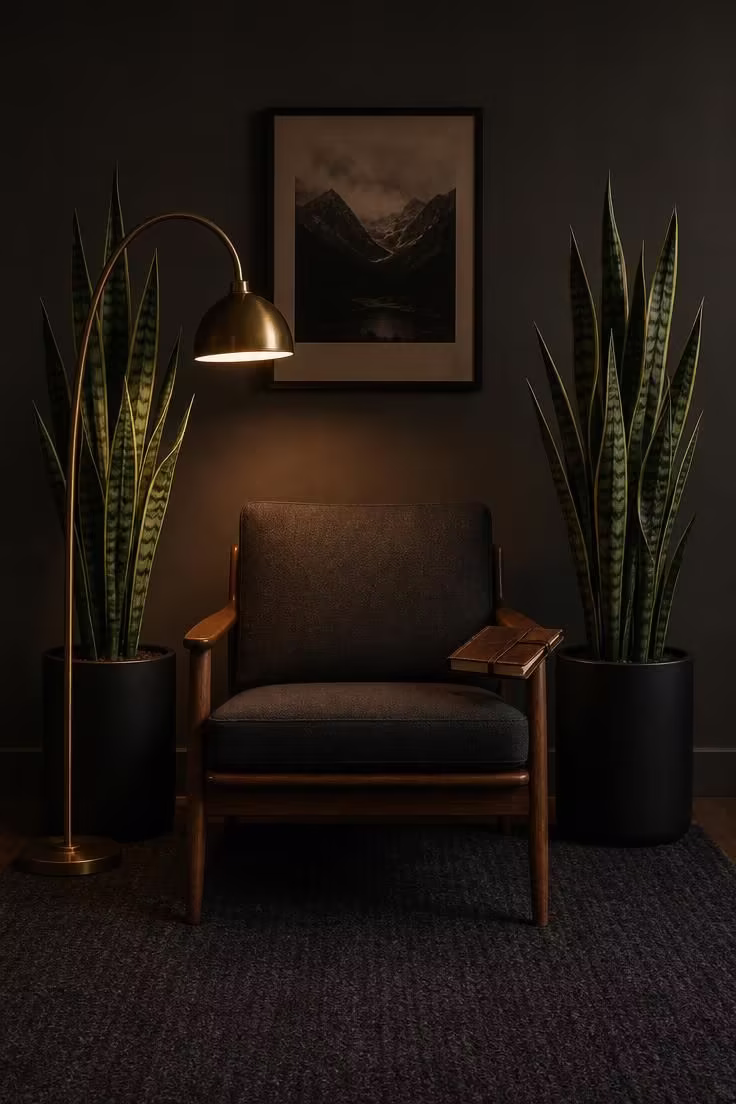 13 Cozy Reading Nook with Plants Ideas You Will Love 11 Two tall snake plants in matte black pots framing a mid-century walnut lounge chair with charcoal wool upholstery under a brass arc lamp in moody low light.