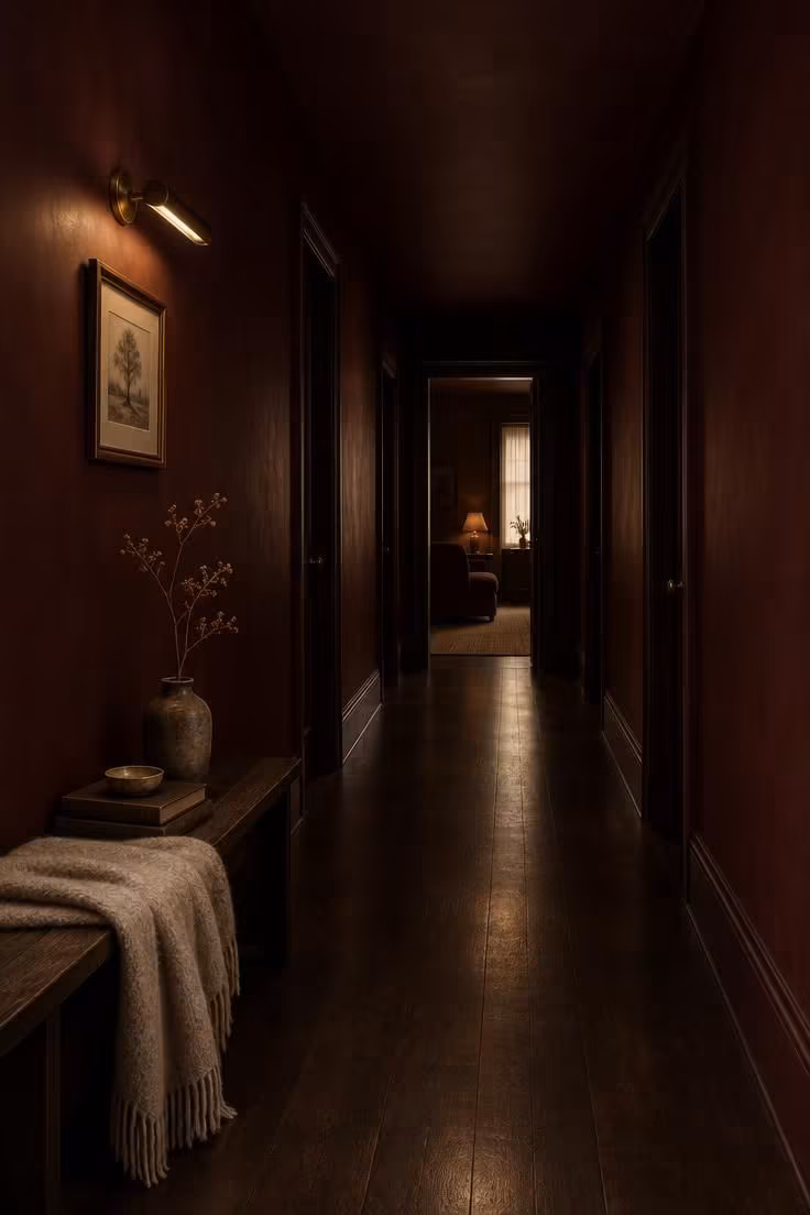 A long narrow corridor with deep oxblood walls and a matching oxblood ceiling, an aged brass picture light, and dark hardwood under a wool-draped bench.