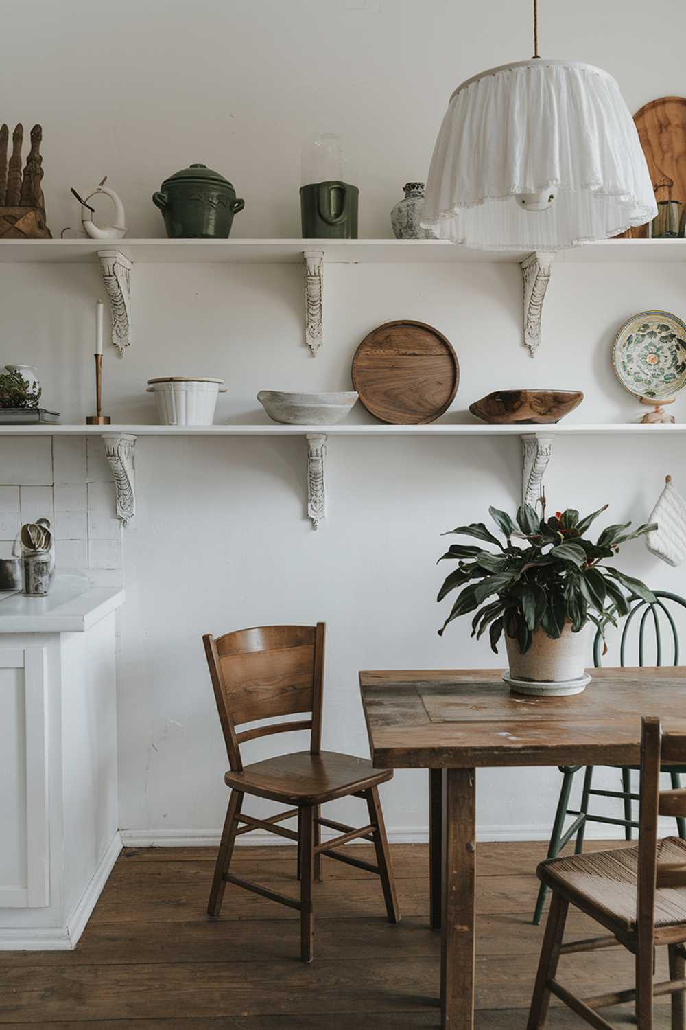 A rustic boho kitchen decor. There's a wooden table with a white lampshade and a potted plant. There are also a few chairs. The walls are covered with white shelves. There are various items on the shelves, including a green pot, a white dish, a wooden dish, and a few decorative items. The floor is made of wood.