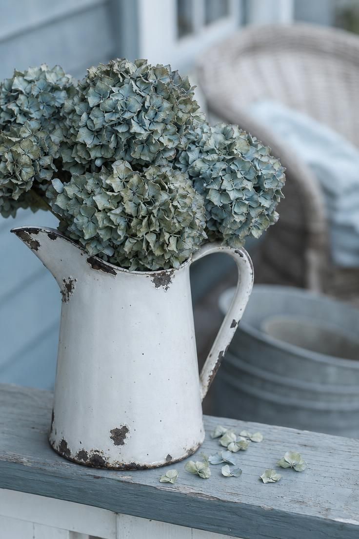 11 Cozy Rustic Fall Porch Ideas for a Warm Welcome 2 A chipped enamel pitcher filled with dried hydrangea blooms on a painted porch railing in soft overcast light.