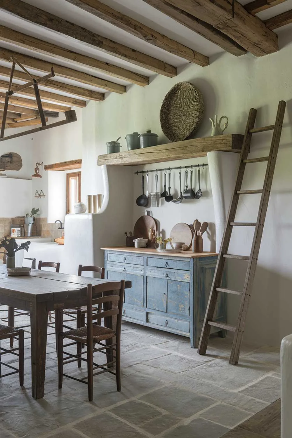 A rustic farmhouse kitchen decor. The kitchen has a wooden beam ceiling, white walls, and a stone floor. There's a large wooden table in the middle of the room with chairs around it. A vintage wooden ladder leans against the wall, holding up a wooden shelf. The shelf has various vintage cooking utensils and a large, round, woven basket. There's a vintage, blue, wooden cabinet against the wall, with a few cooking utensils inside. The room has a rustic, vintage charm.