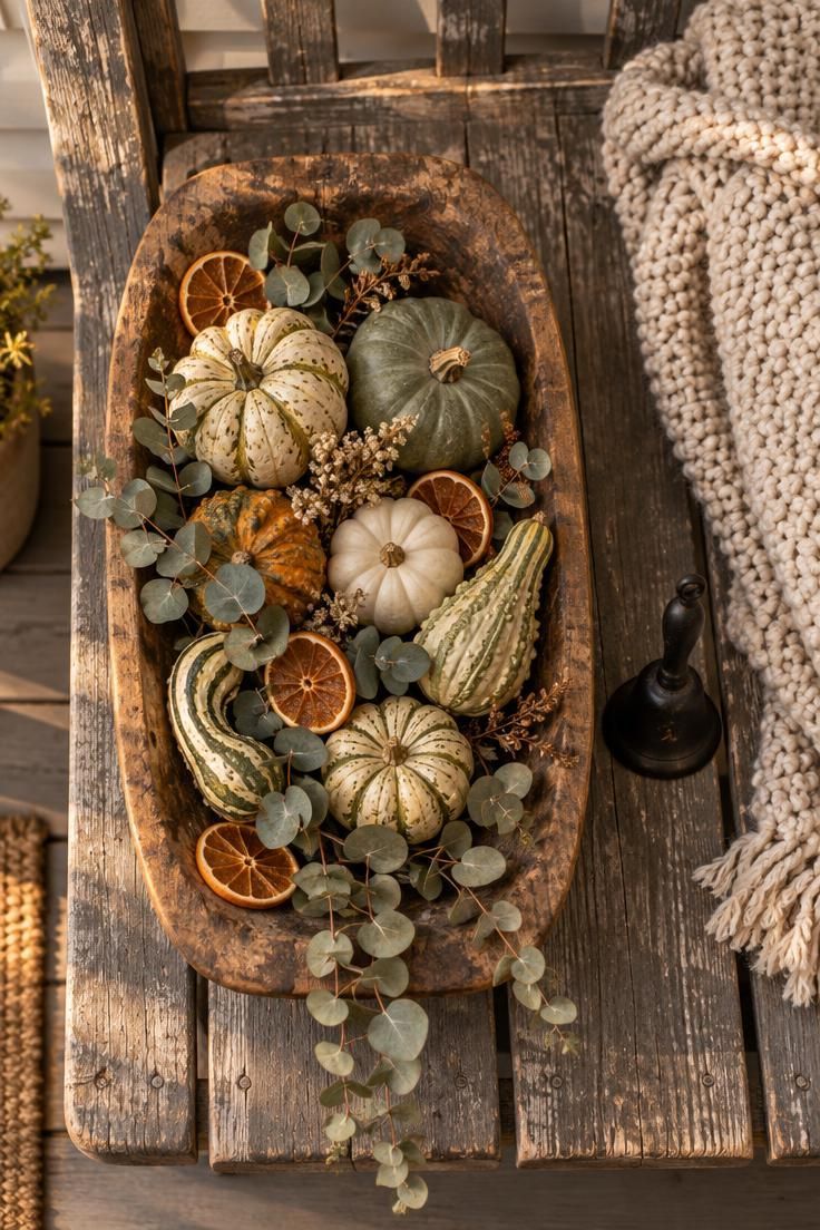 10 Rustic Halloween Decor Ideas for a Welcoming Porch 9 Overhead view of a wooden dough bowl on a porch bench holding sage, cream, and amber gourds with dried orange slices and eucalyptus spilling over the edge.