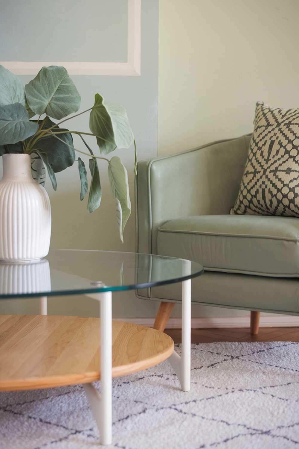 A living room decor with sage green accents. There's a sage green armchair with a white frame and a patterned cushion. Next to the armchair is a white coffee table with a glass top and a wooden base. On the coffee table, there's a white vase with a sage green plant. The floor is covered with a white rug. The background wall has a pale blue hue.