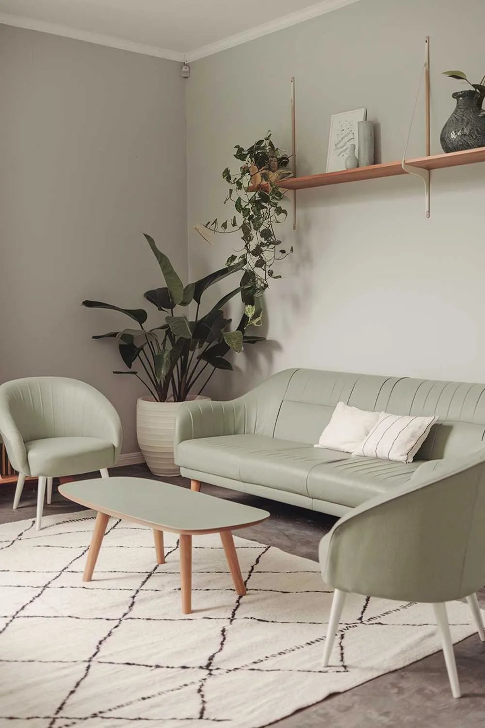 A cozy Scandinavian living room with a sage green color palette. There's a large sofa in sage green with white cushions. A wooden coffee table with a white top sits in front of the sofa. Two sage green chairs with white legs sit near the wall. A wooden shelf with a few decorative items hangs above the sofa. A large potted plant in a white pot is placed near the shelf. The floor is covered with a white rug. The walls are painted a light gray. The lighting is soft.