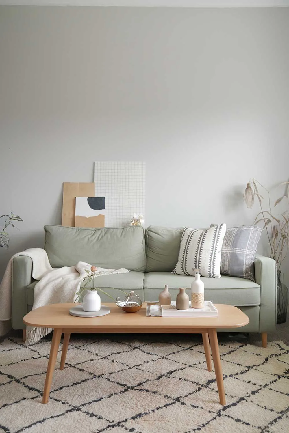 A Scandinavian living room with a sage green couch and a beige rug. There's a wooden coffee table in the middle. There are some decorative items on the coffee table and the wall behind the couch. The walls are painted in a light gray color.